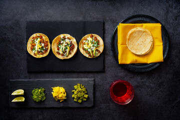 Delicious lunch with tacos and sauces on black table, top view of appetizing Mexican tacos served on black plate near bowl with tomato sauce and avocado placed on wooden table
