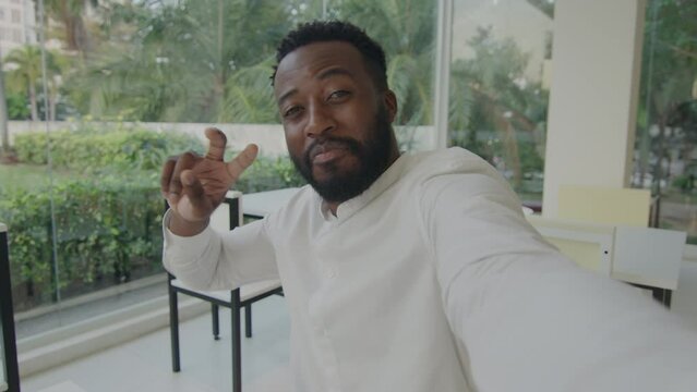 African american blogger man in white shirt in a cafe holding his phone, greeting the audience, making various hand gestures. Chest-up handheld shot with green background