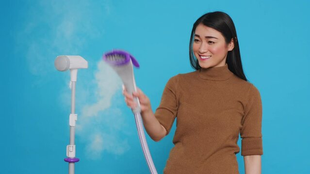 Smiling cleaning lady using steamer to ironing clothes in studio over blue background. Woman used the best cleaning products and equipment to ensure a sparkling, fresh-smelling home for her clients.
