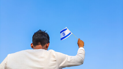Boy holding Israel flag against clear blue sky. Man hand waving Israeli flag view from back, copy space