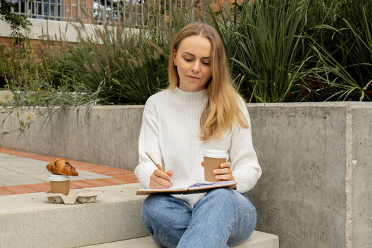 Young Student Study With Notebook In Park. Drinking Coffee And Eat Croissants. Writing Gratitude Journal Self Reflection Self Discovery Outdoors Warm Autumn Seashore. 