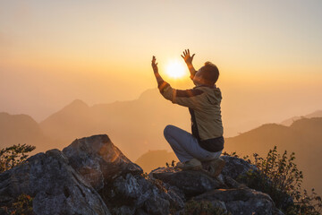 Young male kneeling down with hands open palm up praying to God on the mountain sunset background.