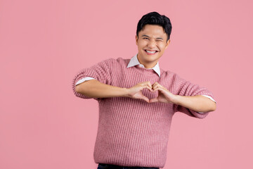 Young asian man wearing casual pink sweater smiling in love doing heart symbol shape with hands....