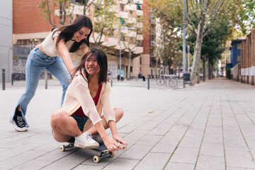 two young women laughing and having a good time playing with a skateboard down a city street,...