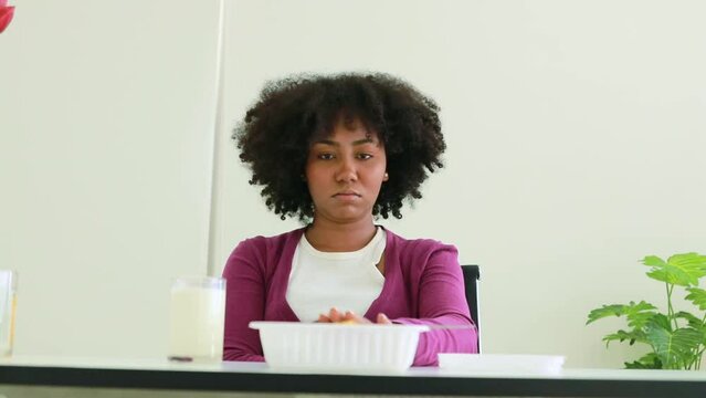 Unhappy young african american woman tired of consuming food packed in plastic containers bought from convenience store, refuses to eat and picks up a glass of milk, gets up from chair and walks away.