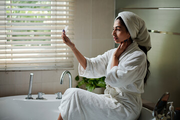 Young woman sitting on edge of bathtub and looking at compact mirror