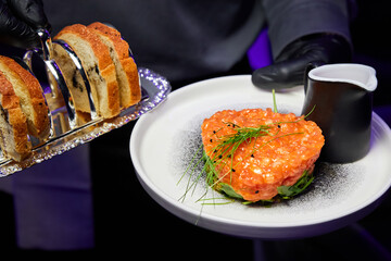 The waiter is holding Salmon tartare with avocado on a white plate in a restaurant. Close-up, selective focus