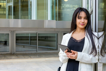 Close up portrait of a young executive businesswoman leaning on glass wall with reflections. Smiling empowered smart casual woman holding a tablet outside the office work site. Horizontal copy space.