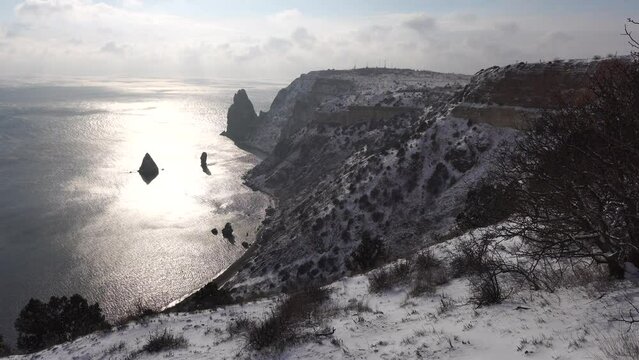 Snow covered rocky cliffs over sea. Winter landscape over the sea bay. Juniper tree under snow. Concept of winter holidays, travel and recreation. Cape Fiolent in Balaklava, Sevastopol, Crimea