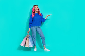 Full length cadre of young funny shopping addicted girl hold palm demonstrate new clothes brand offer with bargains isolated on cyan color background