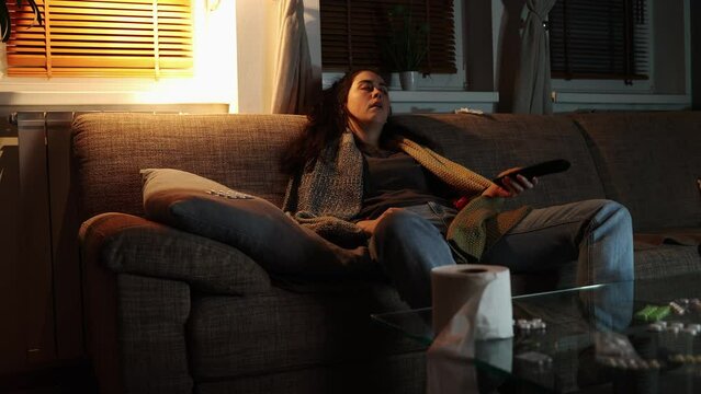 Flu and fever. Illness young Caucasian woman sitting on sofa covered with blanket and watching tv. Home evening. Concept of depression and health problem.