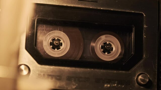 Audio cassette playback in a vintage tape recorder. Record player playing old transparent audio cassette in the flickering light, close-up. Retro tape reels rotate in deck. Record calls, archive, 80s