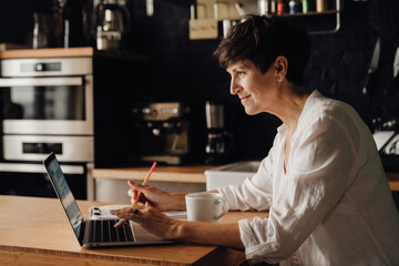 Fototapeta premium Mature woman working on laptop computer while sitting in kitchen at home