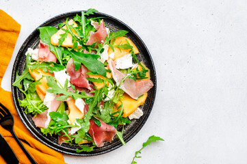 Gourmet lunch melon salad with cantaloupe, prosciutto, soft white cheese and arugula on gray table...
