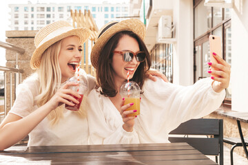 Two young beautiful smiling hipster female in trendy summer straw hats. Sexy carefree women holding and drinking fresh vegetable cocktail smoothie drink in plastic cup with straw. Taking selfie
