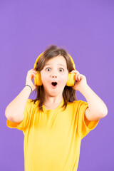 Surprised little girl wearing headphones listening music or hearing podcast isolated on purple background with copy space