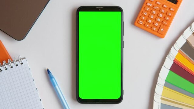 Top view of zoom Mobile Phone Green Screen on Designer Worktop. Close Up. Color Guide, Palette, Laptop, Pen. Cell Phone Mockup with Blank Chromakey Display on a Workplace. Smartphone Layout Color key