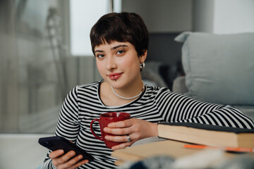 Smiling woman using mobile phone and drinking coffee while sitting on floor in living room