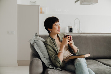 Cheerful positive woman drinking coffee and reading book while sitting on couch