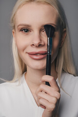 Obraz premium Portrait of a woman covering one eye on the face using makeup brush, looking at camera and smiling isolated over gray.