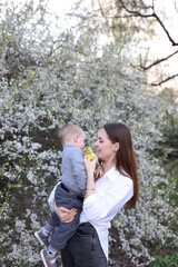 a mother and a little son are sitting on the green grass near a blooming tree and little yellow ducklings are near them. two little ducklings and a one-year-old boy