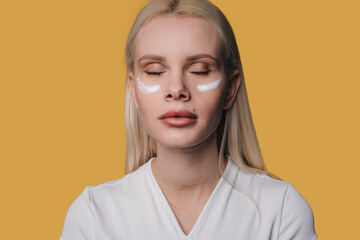 Fototapeta na wymiar Portrait of woman with eyes colesd having healthy face applying cosmetic cream under the eyes isolated over yellow background. Skin care routine. Beauty
