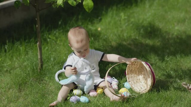 Easter holiday outdoor with baby child kid. Toddler baby bunny ears playing Easter eggs pouring them out of basket falling on green grass. Spring activities children outdoors Easter games