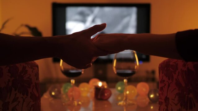 Lovers holding hands watching movie in living room. Hands of young couple close up. Happy couple of lovers hold each other's hands. Anniversary celebration at home.