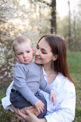 Fototapeta na wymiar mother hugs her little son in spring near a blooming tree. Spring and plum blossom. Mother's Day. The love of a son and a mother. Gray clothes for a one-year-old boy. Kiss the son. child's laughter