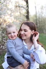 Fototapeta na wymiar mother hugs her little son in spring near a blooming tree. Spring and plum blossom. Mother's Day. The love of a son and a mother. Gray clothes for a one-year-old boy. Kiss the son. child's laughter