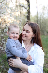 Fototapeta premium mother hugs her little son in spring near a blooming tree. Spring and plum blossom. Mother's Day. The love of a son and a mother. Gray clothes for a one-year-old boy. Kiss the son. child's laughter