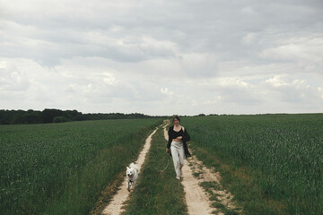 Fototapeta na wymiar Woman walking with cute white dog in wheat field. Stylish young female hiking and running with funny dog in summer countryside. Pet and travel. Danish spitz