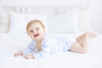 laughing baby blond boy lies in a crib on his stomach in a room at home with blue and white cotton bedding and smiles in the morning, the concept of children's goods and accessories