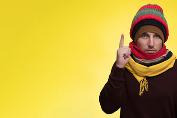 A man on a yellow background in several hats and scarves holds up an index finger.