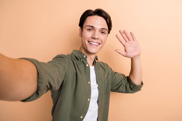 Photo of optimistic handsome cool guy with brunet hair dressed khaki shirt doing selfie waving palm isolated on beige color background