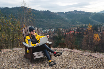 Woman wearing glasses and yellow jacket working on laptop outdoors surrounded by beautiful nature,...