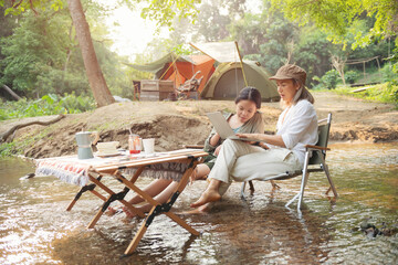 Pleased happy mother and daughter reading a book and using laptop while relaxing on the deck chairs in the river, sit near a camp and tent, drink coffee in a pine forest. Camping, recreation, hiking.