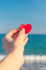 Fototapeta premium The red heart in the hand of the girl against the background of the sea horizon during sunset.