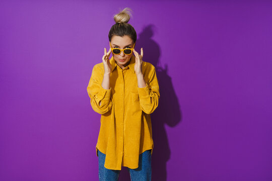 Young  girl wearing yellow shirt and sunglasses making silly face expression against purple background