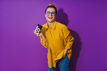 Fototapeta na wymiar Cheerful girl wearing eyeglasses holding vintage film camera on purple background