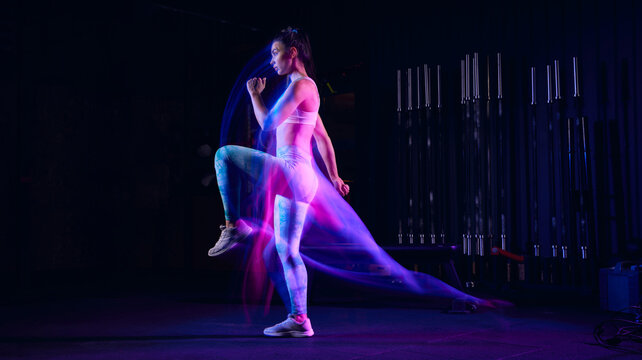 Workout at gym. Sportive woman in sportswear and sportsfoot training in modern fitness room over dark background with mixed neon light. Active and healthy lifestyle