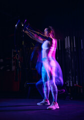 Portrait of muscular strong sportive young girl doing exercises, workout at dark gym in mixed neon light. Active and healthy lifestyle, sport, fitness concept