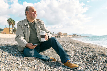Happy middle-aged bearded man in casual clothes using smartphone and drinking coffee while while sitting on winter on spring beach. Relaxing outdoors.
