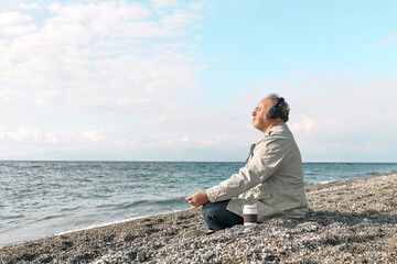 Naklejka na ściany i meble Happy middle-aged bearded man sitting in lotus pose on pebble winter beach, meditating and listening music or podcast from smartphone application in headphones. Relaxing outdoors.