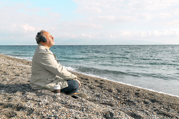 Happy middle-aged bearded man sitting in lotus pose on pebble winter beach, meditating and...