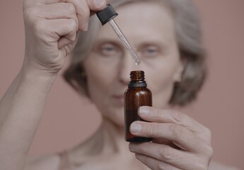 CU Portrait of beautiful grey-haired 60s mature senior adult female holding brown amber glass cosmetic liquid bottle without label, product mockup. Studio shot, soft lighting