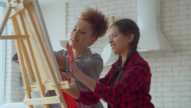 Happy creative cute multiracial adolescent daughter and positive charming black mother spending leisure together, having fun painting with hands on easel using colorful paints at home.