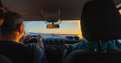 A middle-aged couple driving a 4x4 from inside the car at dawn on a totally snowy road with the sun rising on the horizon and the driver's gaze reflected in the rearview mirror