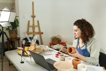 Females potter in blue apron making a vlog, recording online course, clay master class,lessons in her studion with earthenware.Using led lamp,studio equipment