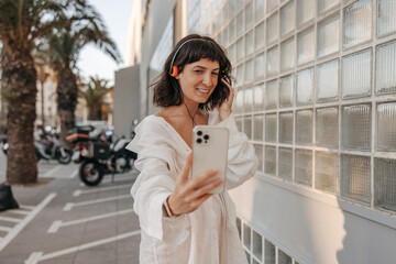Obraz premium Charming fair-skinned young woman doing selfie in phone at the street. In headphones, look with smile on the street background. Positive emotion, technology concept 
